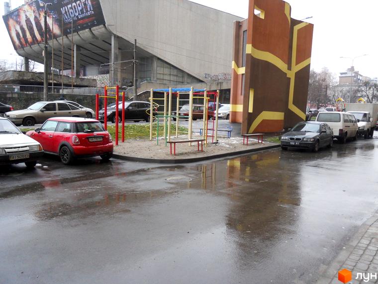 Велика сіра будівля зі схилим дахом та рекламним плакатом, поруч коричнева споруда з жовтими візерунками. Перед ними дитячий майданчик та мокра дорога з автомобілями.