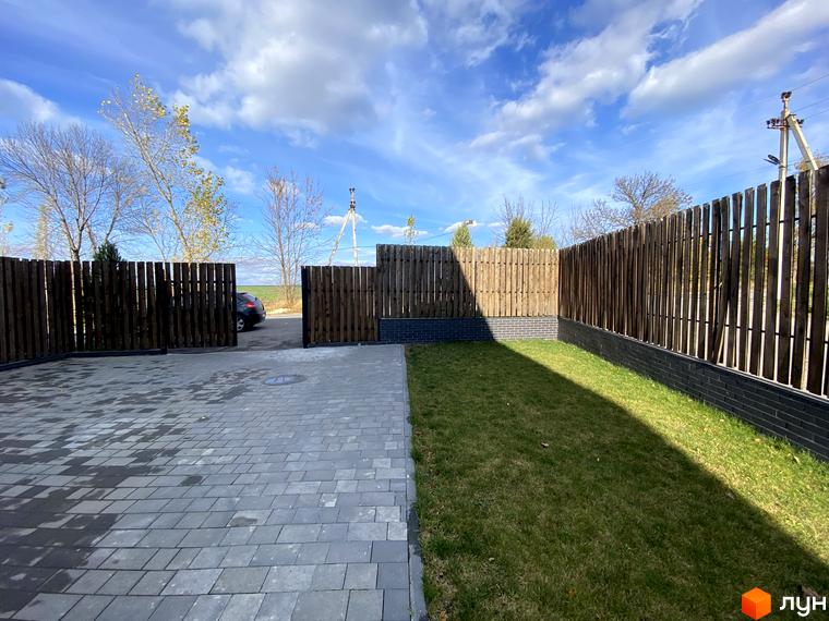 Таунхаус „Новоолександрівський“: дерев'яний паркан, вимощена сіра плитка та зелений газон. Огляд прибудинкової території та парковки біля будинку.