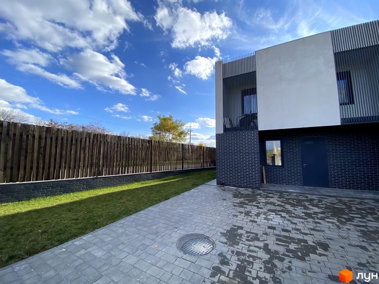 Таунхаус Новоолександрівський з цегляним фасадом та сірою бруківкою на прибудинковій території. Будинок має два поверхи з білим оздобленням та вертикальними елементами.