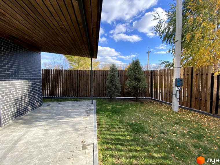 Таунхаус „Новоолександрівський“ з сірою цегляною стіною, дерев’яним навісом та вимощеною плиткою терасою. Поруч зелений газон та дерев'яний паркан.