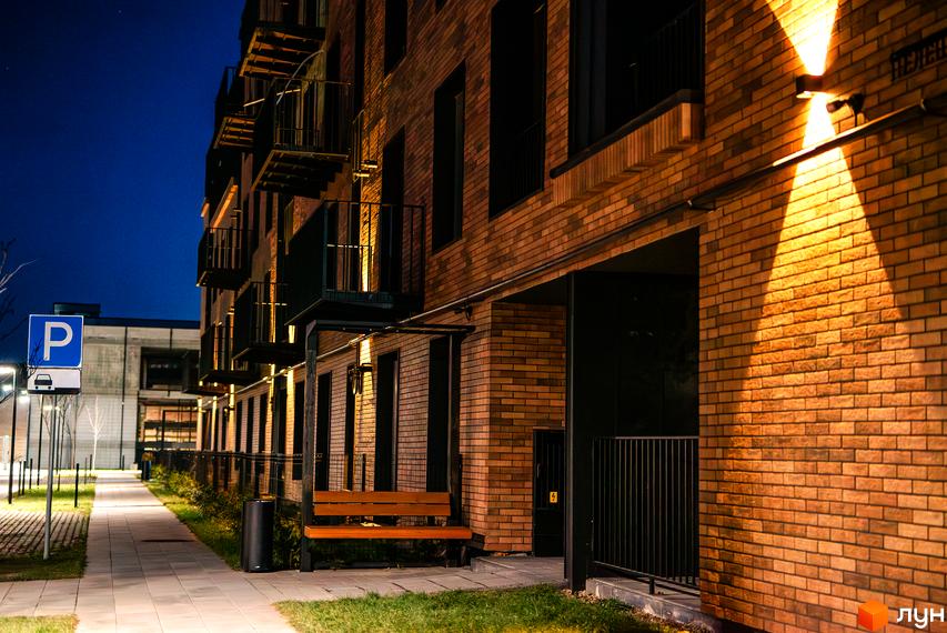 Зовнішній вигляд будинку ЖК Клубне містечко 12 з цегляним фасадом та балконами, освітлений вночі. Поруч розташована доглянута прибудинкова територія з лавкою та парковою зоною.