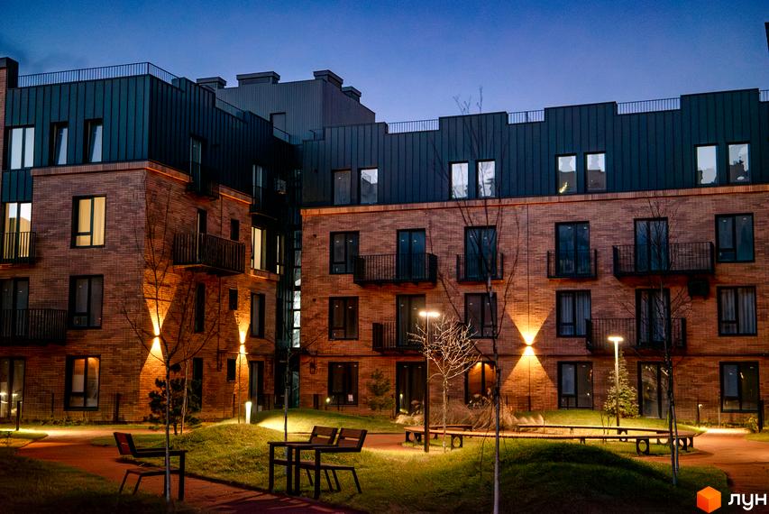 Клубне містечко 12. Фасад будинку з цегляною обробкою та темною металевою покрівлею, освітлена прибудинкова територія з ландшафтним дизайном та місцями для відпочинку.