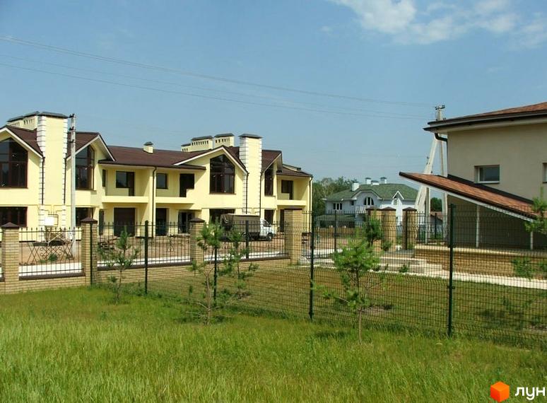 Зображення житлового комплексу з будинками у світлих тонах з коричневими дахами. На передньому плані зелена трава та огорожа.