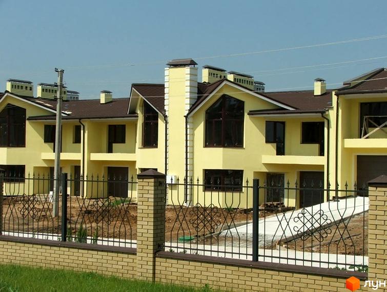 Житловий комплекс з жовтими будинками та коричневими дахами. На передньому плані цегляний паркан з кованими елементами.