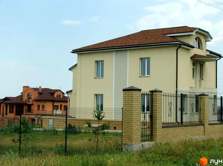 Двоповерховий будинок з бежевими стінами та коричневим дахом у Котеджному містечку Молодежная резиденция. Перед будинком розташований газон з невисокою травою та металевий паркан.