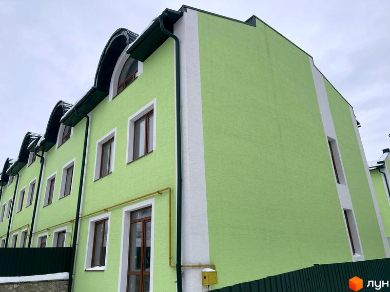 Таунхауси „Гаї Гречинські“: двоповерхові будинки світло-зеленого кольору з білими вертикальними смугами та темними дахами. Навколо будівель видно зелений паркан та елементи зимового пейзажу.