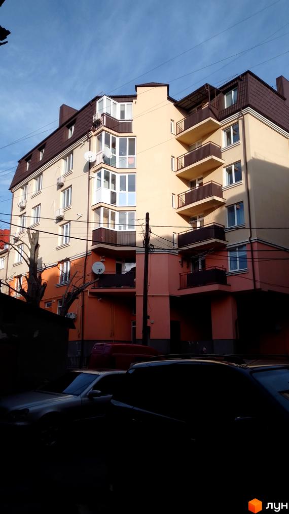 Багатоповерховий будинок з балконами та еркерами, пофарбований у світлі тони. На передньому плані припарковані автомобілі та дерева.