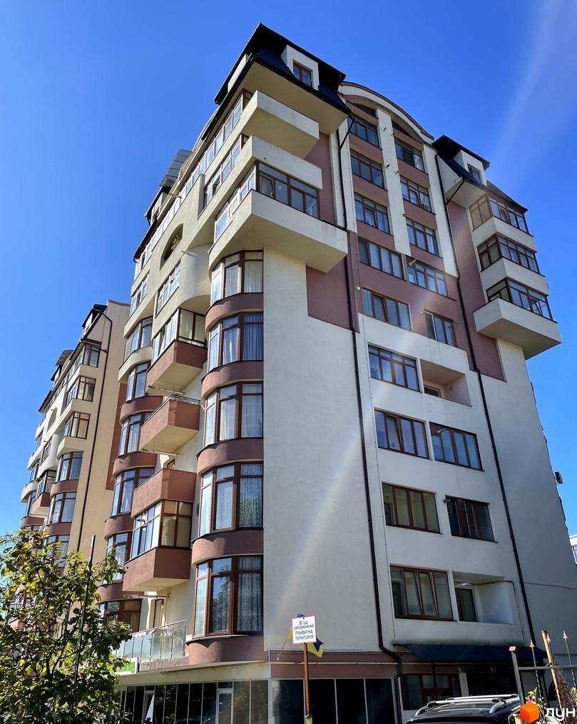 Висотна будівля житлового комплексу з великою кількістю вікон та балконів. Фасад будинку пофарбовано у світлі тони з темними вставками.