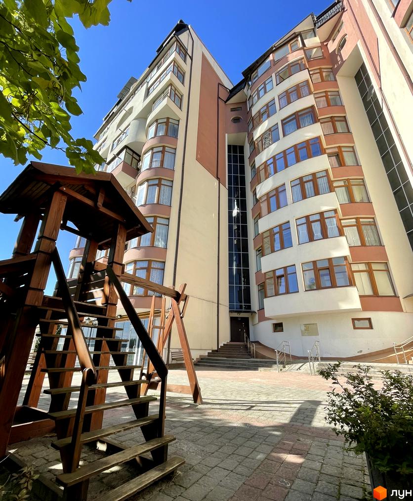 Висотна будівля житлового комплексу з великими вікнами та балконами. На передньому плані дитячий майданчик з дерев'яними конструкціями.