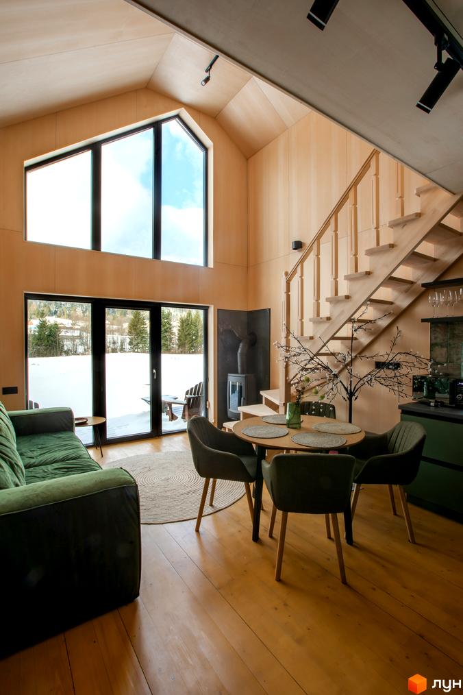 Інтер'єр котеджного містечка TYSHA з великими вікнами, дерев'яними сходами та каміном. З вікон відкривається вид на засніжений пейзаж з деревами.