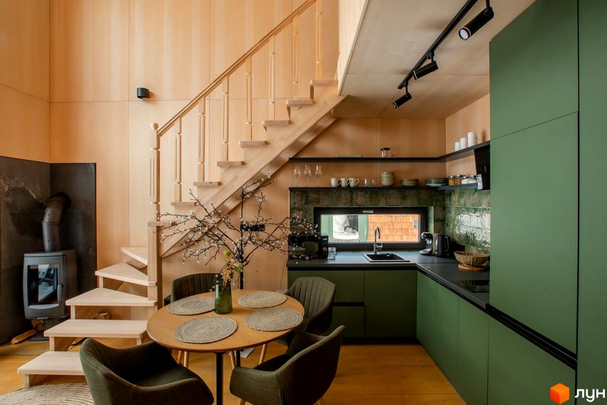 Інтер'єр кухні та вітальні з каміном та дерев'яними сходами в котеджі КМ TYSHA. Простора кухня з зеленими фасадами та обідня зона з круглим столом.