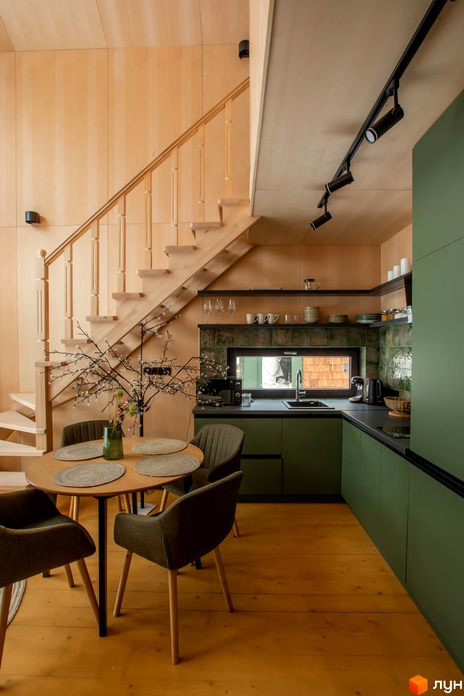 Інтер'єр кухні-вітальні з дерев'яними сходами та зеленими кухонними фасадами. Затишна обідня зона з круглим столом та кріслами.