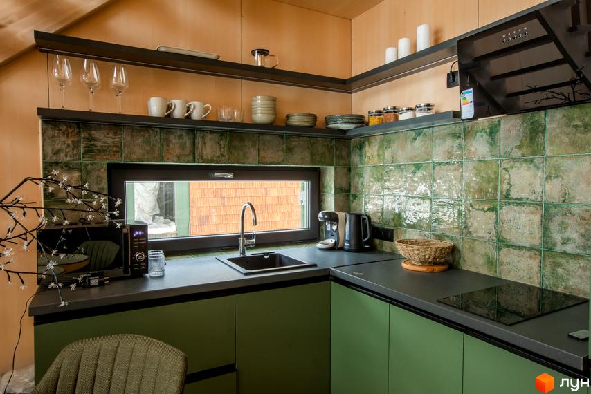 Сучасна кухня з зеленими кахельними стінами та дерев'яними елементами. Кухонні меблі виконані в зеленому кольорі, стільниця темна, є вбудована техніка.