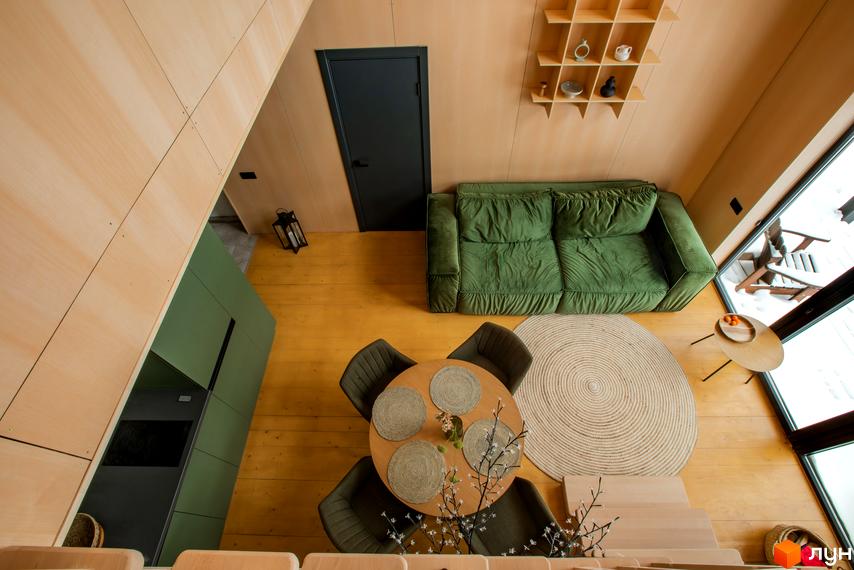 Інтер'єр котеджного містечка TYSHA: зелений диван, обідній стіл з кріслами та дерев'яні елементи створюють затишну атмосферу. Велике вікно відкриває вид на засніжену територію.