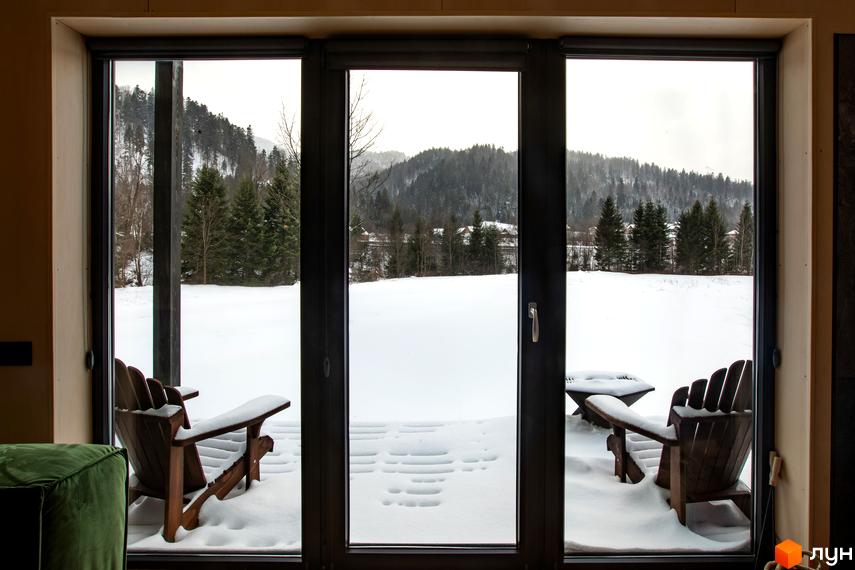 Котеджне містечко TYSHA з дерев'яними кріслами та столом на засніженій терасі. Навколо ліс та гори, що створюють затишну зимову атмосферу.