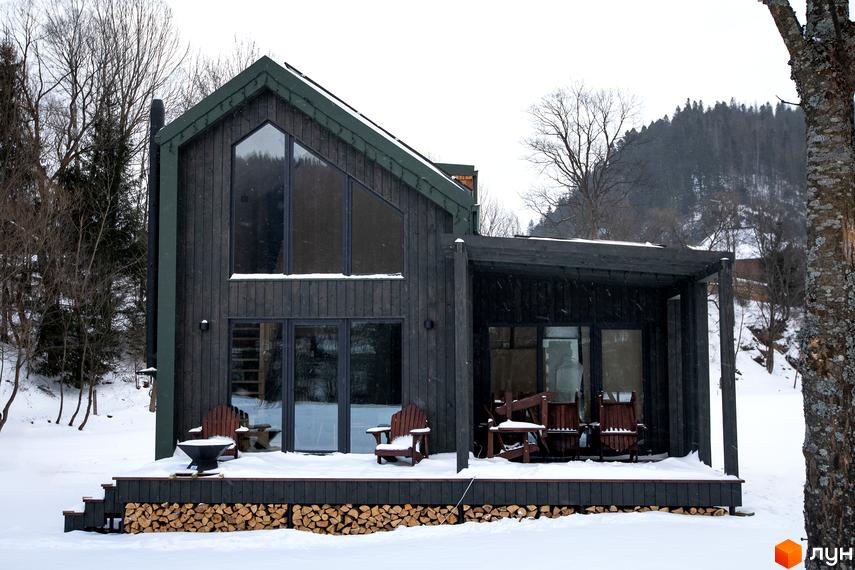 Котеджне містечко TYSHA у Карпатах, будинок з темного дерева та великими вікнами на тлі засніженого лісу. Тераса з дерев'яними кріслами та дровами.