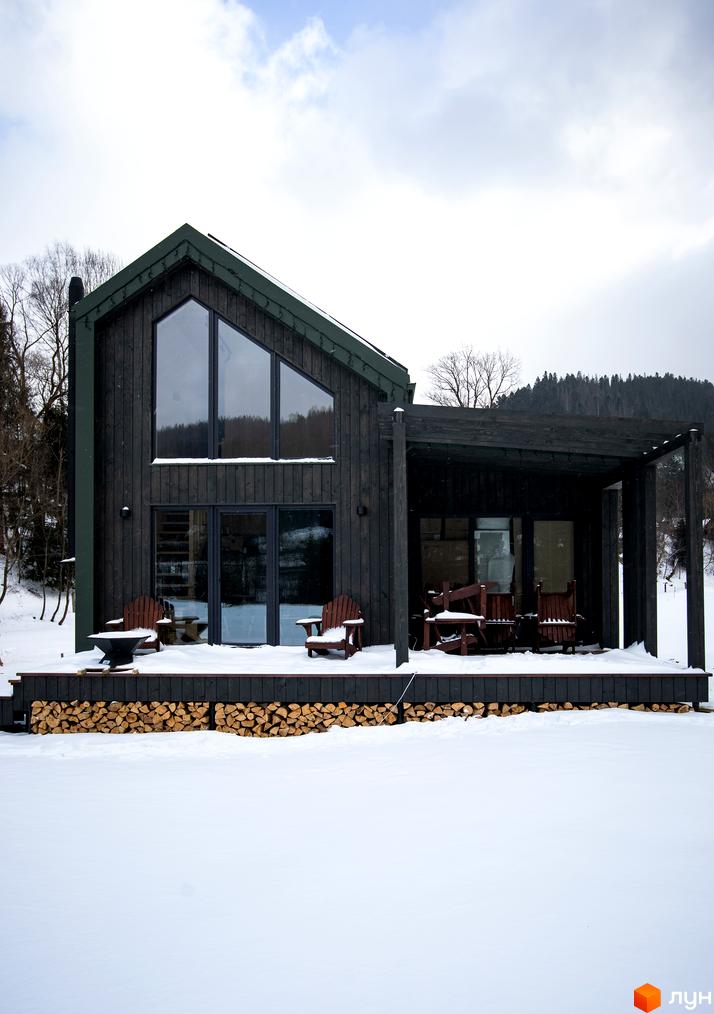 Котеджне містечко TYSHA: дерев'яний будинок з великими вікнами та терасою, оточений зимовим пейзажем та дровами.