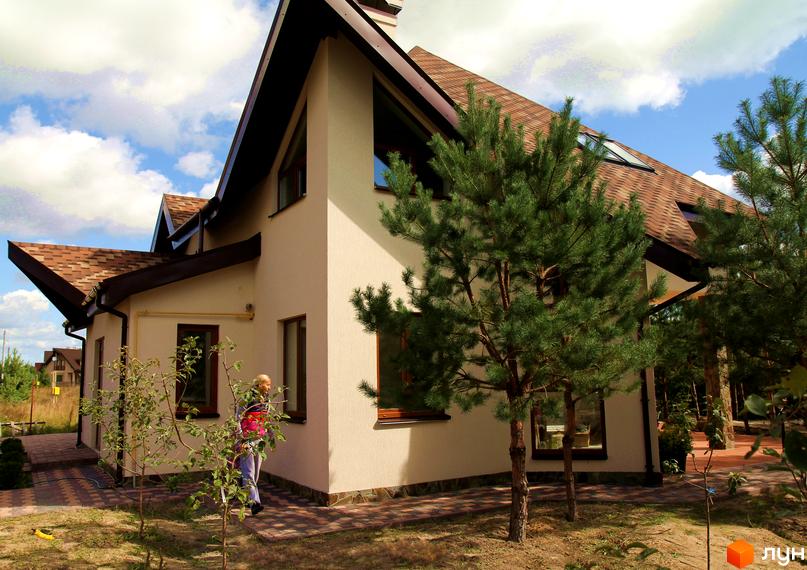 Двоповерховий будинок з коричневим дахом та світлими стінами. На території Котеджне містечко Лелечий Хутір ростуть дерева та кущі.