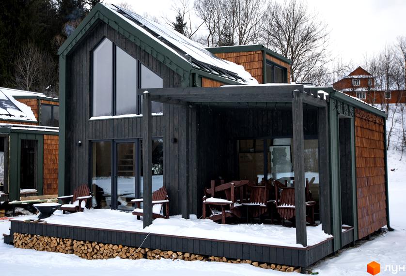 Котеджне містечко КМ TYSHA: дерев'яний будинок з великими вікнами та терасою, оточений зимовим пейзажем з ялинами та снігом.