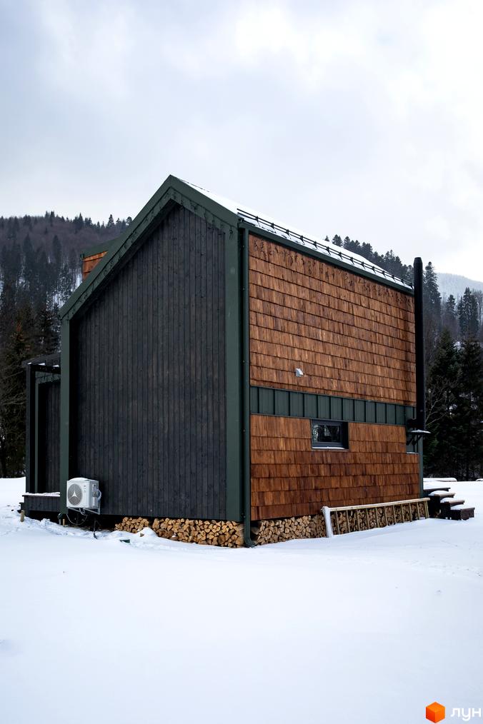 Котеджне містечко TYSHA у Карпатах, дерев'яний будинок з двосхилим дахом та дровітнею на тлі засніженого лісу.