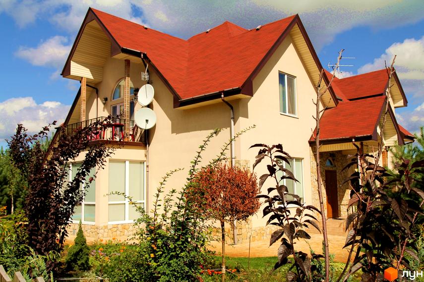 Зображення будинку з червоним дахом та балконом. Перед будинком розташована прибудинкова територія з рослинами та деревами.