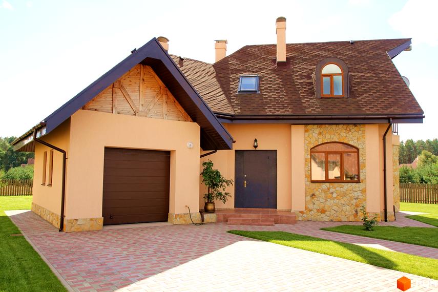 Одноповерховий будинок з коричневою черепичною покрівлею та світло-бежевими стінами. Перед будинком викладена бруківка, є газон та декоративні рослини.