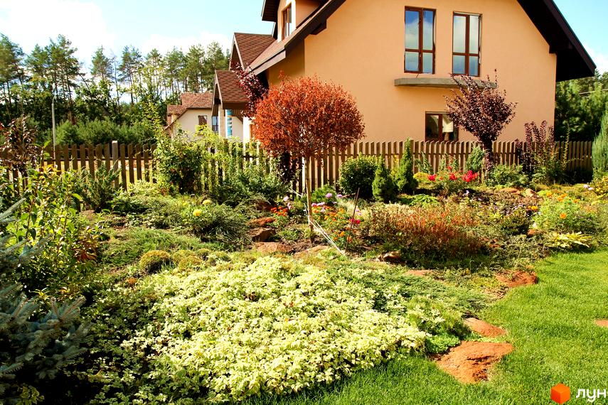Житловий комплекс з теракотовим фасадом та великими вікнами. На передньому плані доглянутий сад з різноманітними рослинами та газоном.