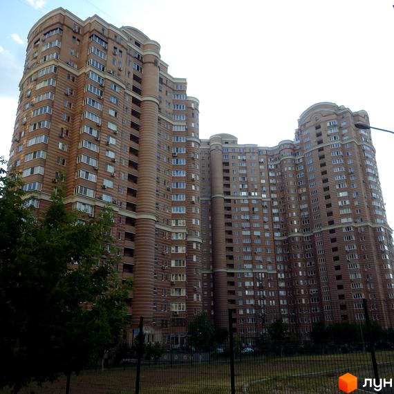 Житловий комплекс Голосіїво: багатоповерховий будинок світло-коричневого кольору з хвилястим фасадом та великою кількістю вікон. Перед ним зелена зона з деревами.