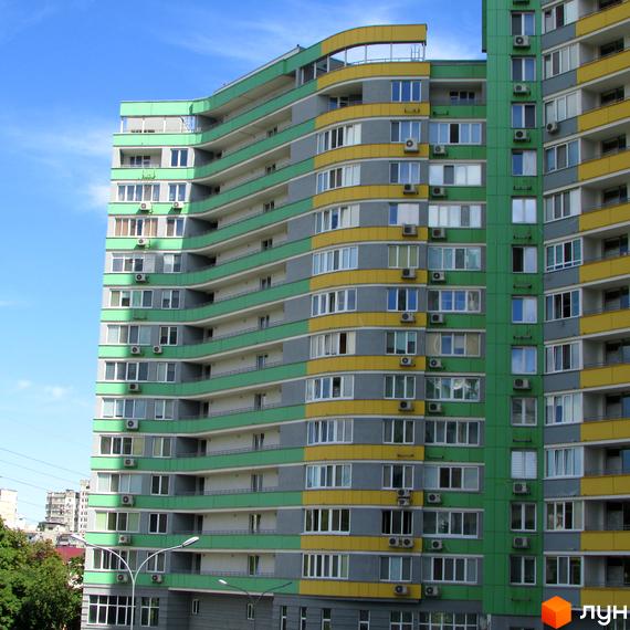 Багатоповерховий житловий комплекс Паркове місто з вигнутим фасадом зеленого, сірого та жовтого кольорів, з численними вікнами та балконами. На тлі блакитного неба та зелені дерев.