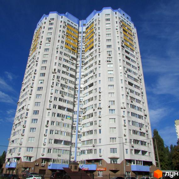 Високий Житловий комплекс Авіатор зі світло-сірим фасадом, синіми та жовтими балконами, та численними вікнами. На першому поверсі розташовані комерційні приміщення, поруч дерева.