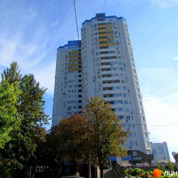 Житловий комплекс Авіатор: високий житловий будинок світло-сірого кольору з синьо-жовтими балконами. На передньому плані зелені дерева.