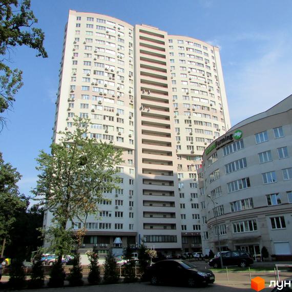 Житловий комплекс Вікторія: високий житловий будинок світлого відтінку з вигнутим фасадом, численними вікнами та кондиціонерами. Поруч зелені насадження та парковка.