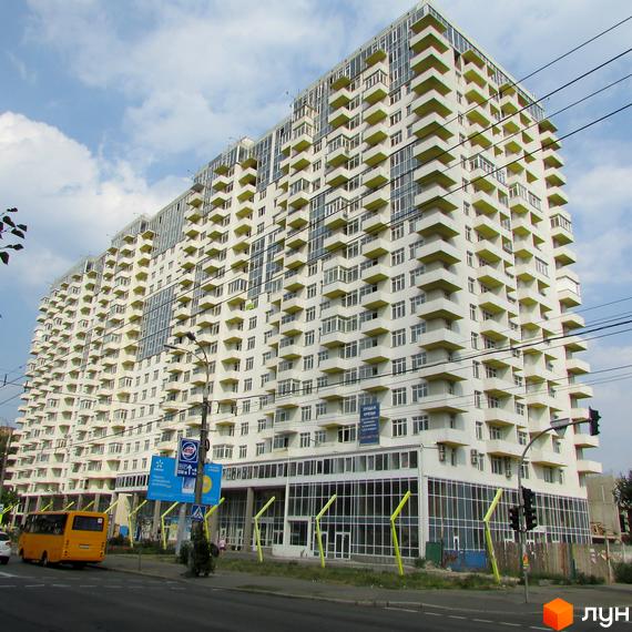 Житловий комплекс Перлина Нивок з білим фасадом та жовтими балконами. Вигляд з вулиці з дорогою, автобусом та дорожніми знаками.