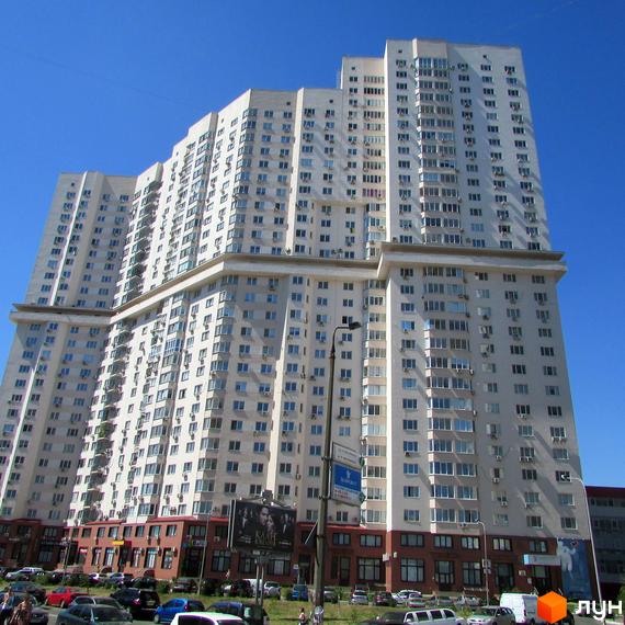 Багатоповерховий житловий будинок світлого кольору зі ступінчастою формою та численними вікнами. Перед будинком розташована парковка.