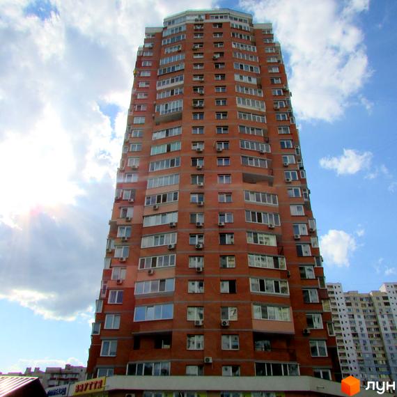 Вид на житловий комплекс з червоної цегли, що має балкони та великі вікна. Прибудинкова територія облаштована для комфортного проживання.