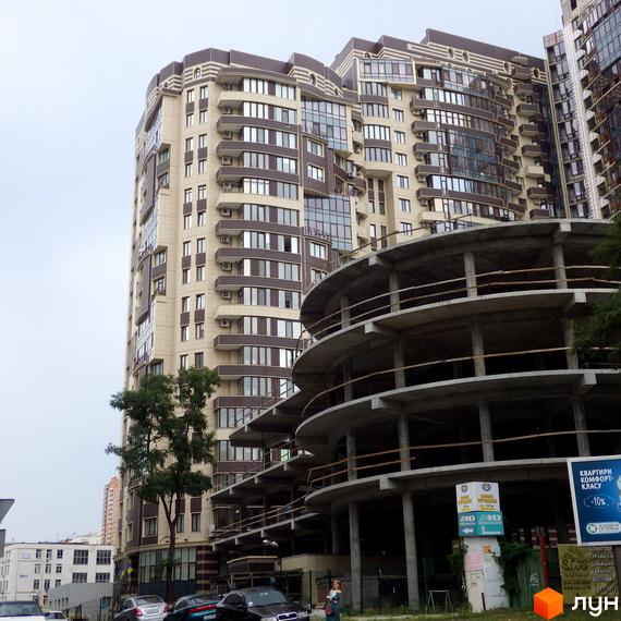 Житловий комплекс Престиж Холл з бежево-коричневим фасадом та численними вікнами. На передньому плані недобудована кругла парковка.