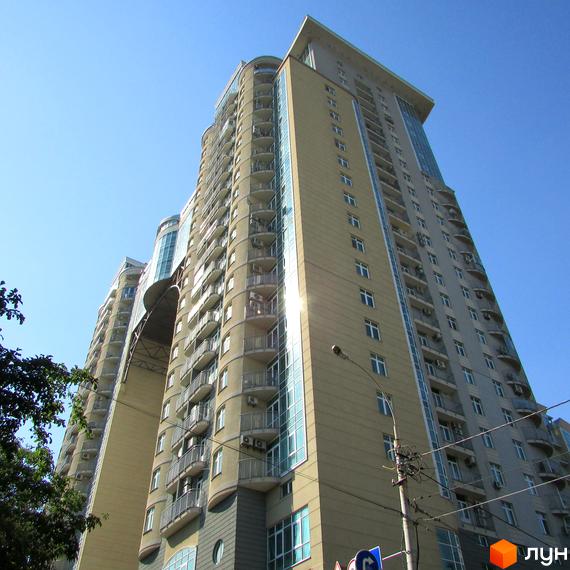 Багатоповерховий житловий комплекс Тріумф з бежевим фасадом, балконами та скляними елементами. Видно блакитне небо та зелень дерев.