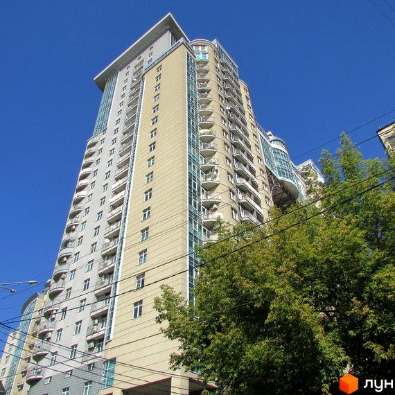 Житловий комплекс Тріумф – багатоповерхова будівля з бежево-сірим фасадом, численними балконами та великими вікнами. Навколо видно зелені дерева та чисте небо.