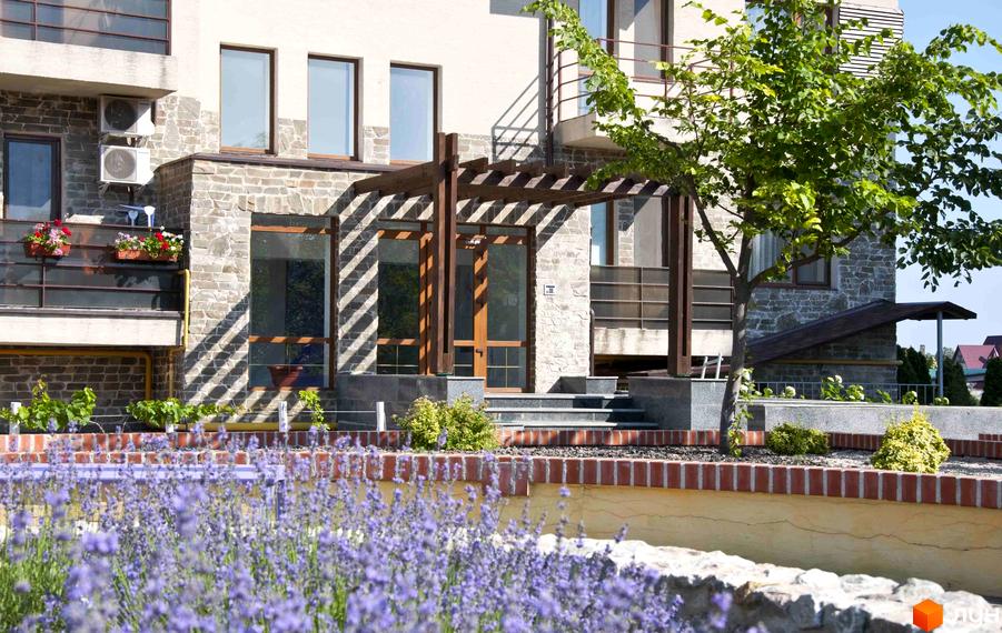 Житловий комплекс Premium Residence (Зелений Мис) з кам'яним фасадом та дерев'яною перголою. Навколо будинку розташовані зелені насадження та квіти.