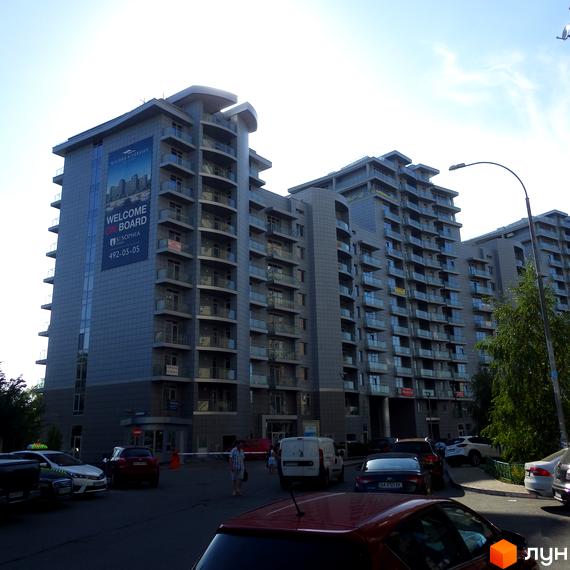 Фасад Житлового комплексу Riviera Riverside з балконами та великим рекламним банером. На передньому плані видно парковку з автомобілями та зелені насадження.