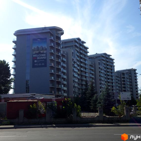 Багатоповерхові будинки Житлового комплексу Рів'єра Ріверсайд з балконами, розташовані вздовж дороги. Перед будівлями видно зелені дерева та огорожу.