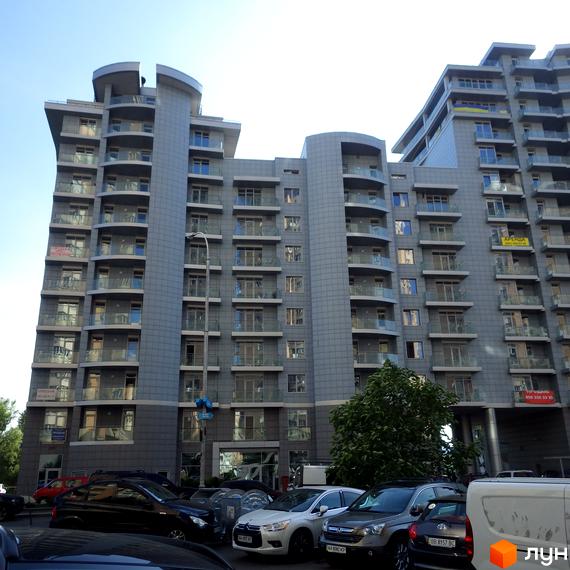 Житловий комплекс Рів'єра Ріверсайд: багатоповерховий будинок з сірим фасадом та балконами зі скляними огорожами. Перед будинком парковка з автомобілями.