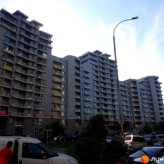 Житловий комплекс Рів'єра Ріверсайд: сірі багатоповерхові будинки з численними балконами, припарковані автомобілі та зелені насадження біля дороги.