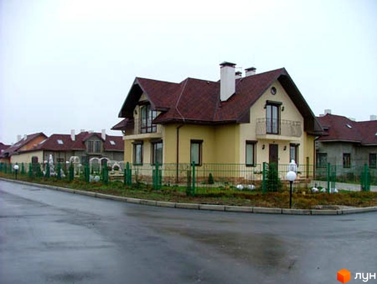 Зовнішній вигляд будинку в Котеджному містечку Петрушки з червоною покрівлею та прибудинковою територією. Видно зелену огорожу та освітлення.