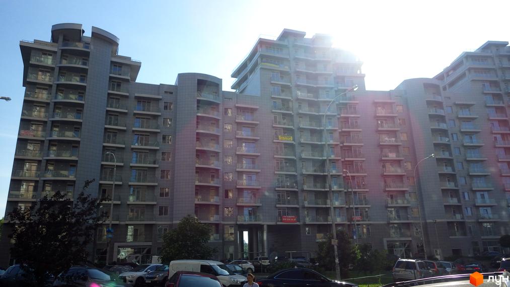 Житловий комплекс Рів'єра Ріверсайд: багатоповерховий будинок з сірим фасадом та балконами. Перед будівлею парковка та зелені насадження.