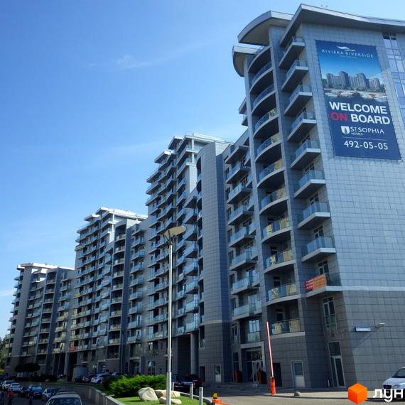 Житловий комплекс Riviera Riverside: багатоквартирні будинки з багатьма балконами та скляними огорожами. Прибудинкова територія з парковкою та зеленою зоною.