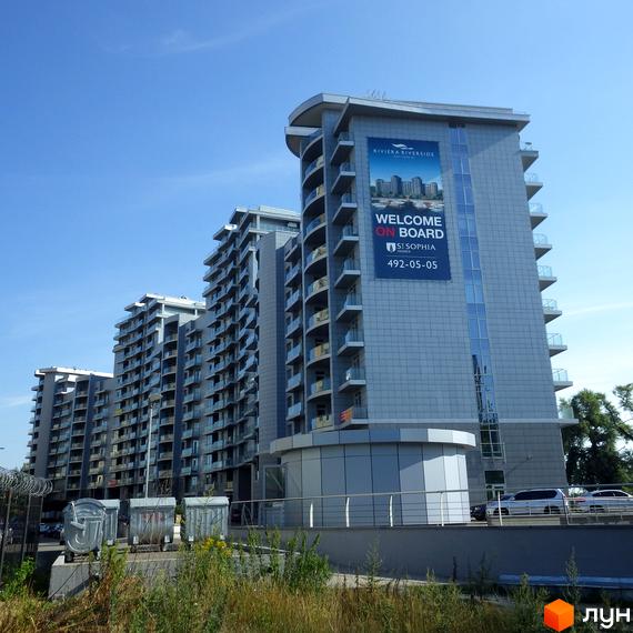 Багатоповерхові будинки житлового комплексу Riviera Riverside з сірими фасадами та скляними балконами. На передньому плані видно зелену траву.