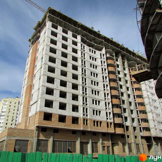 Будівництво Житлового комплексу Перлина Кадетського Гаю: багатоповерховий будинок зі світлих блоків та червоної цегли, з вигнутим фасадом та балконами. На передньому плані зелений паркан.