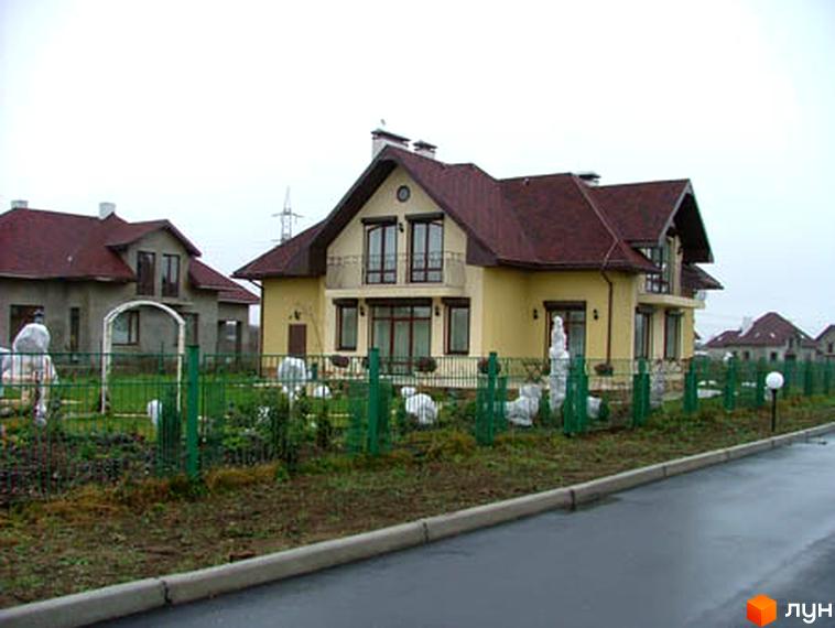 Зображення житлового будинку в Котеджному містечку Петрушки. На фото видно двоповерховий будинок з коричневою черепицею та жовтими стінами, а також зелений паркан навколо території.