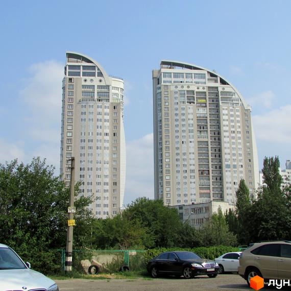 Зображення двох високих житлових будинків світлого кольору з вигнутими дахами, оточених зеленню та припаркованими автомобілями.